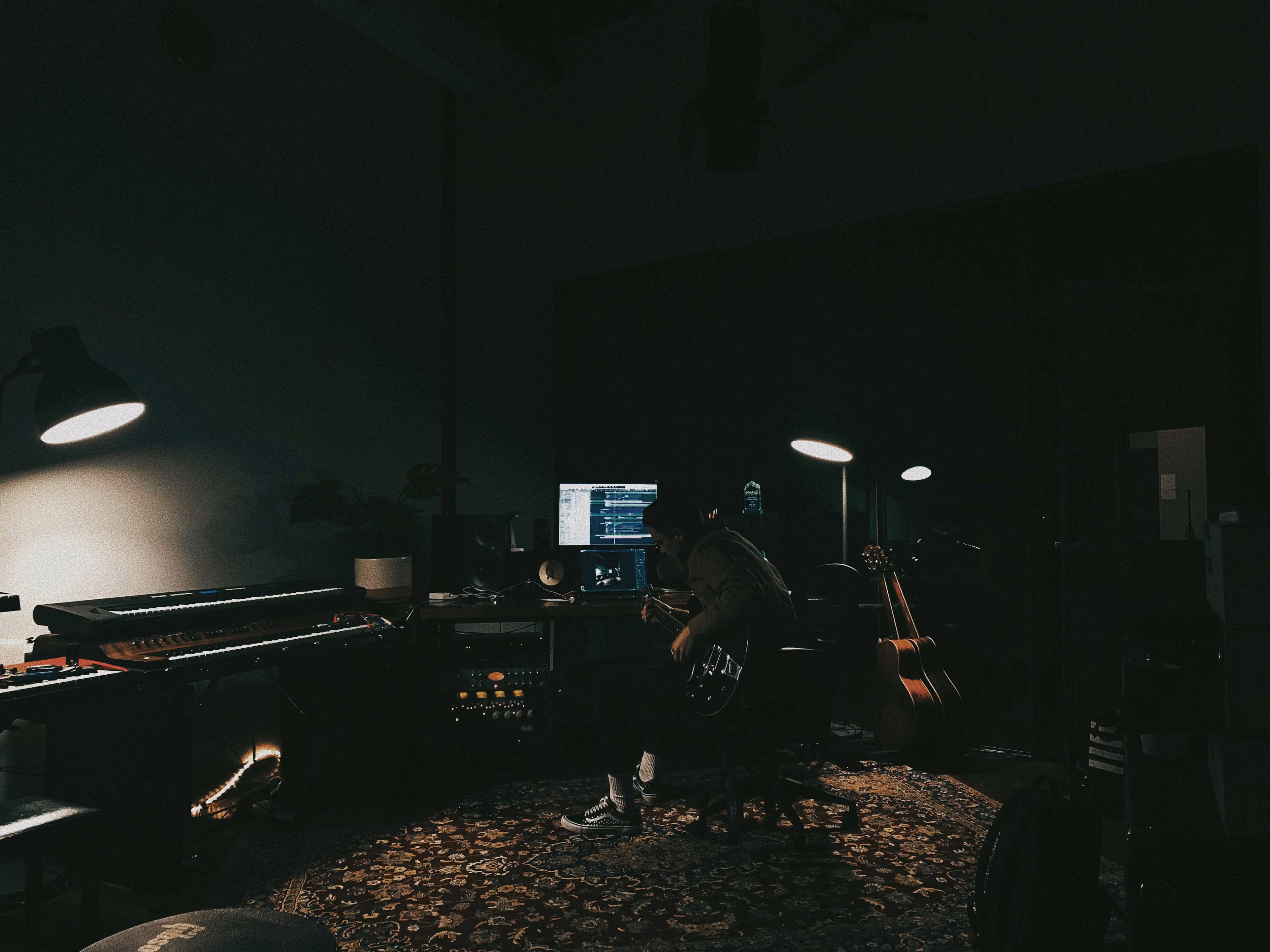 A songwriter playing guitar at night in a dimly lit recording studio, surrounded by keyboards and studio monitors